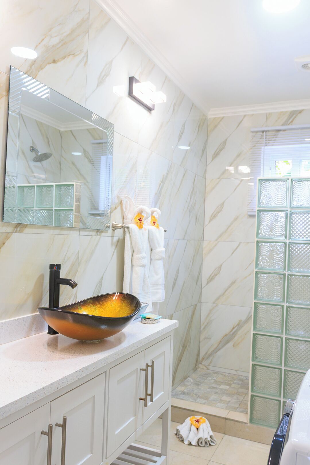Un baño con pared de mármol, un espejo grande, un lavabo sobre encimera, armarios blancos y una zona de ducha.