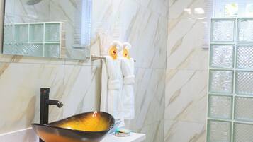 Un baño con pared de mármol, un espejo grande, un lavabo sobre encimera, armarios blancos y una zona de ducha.