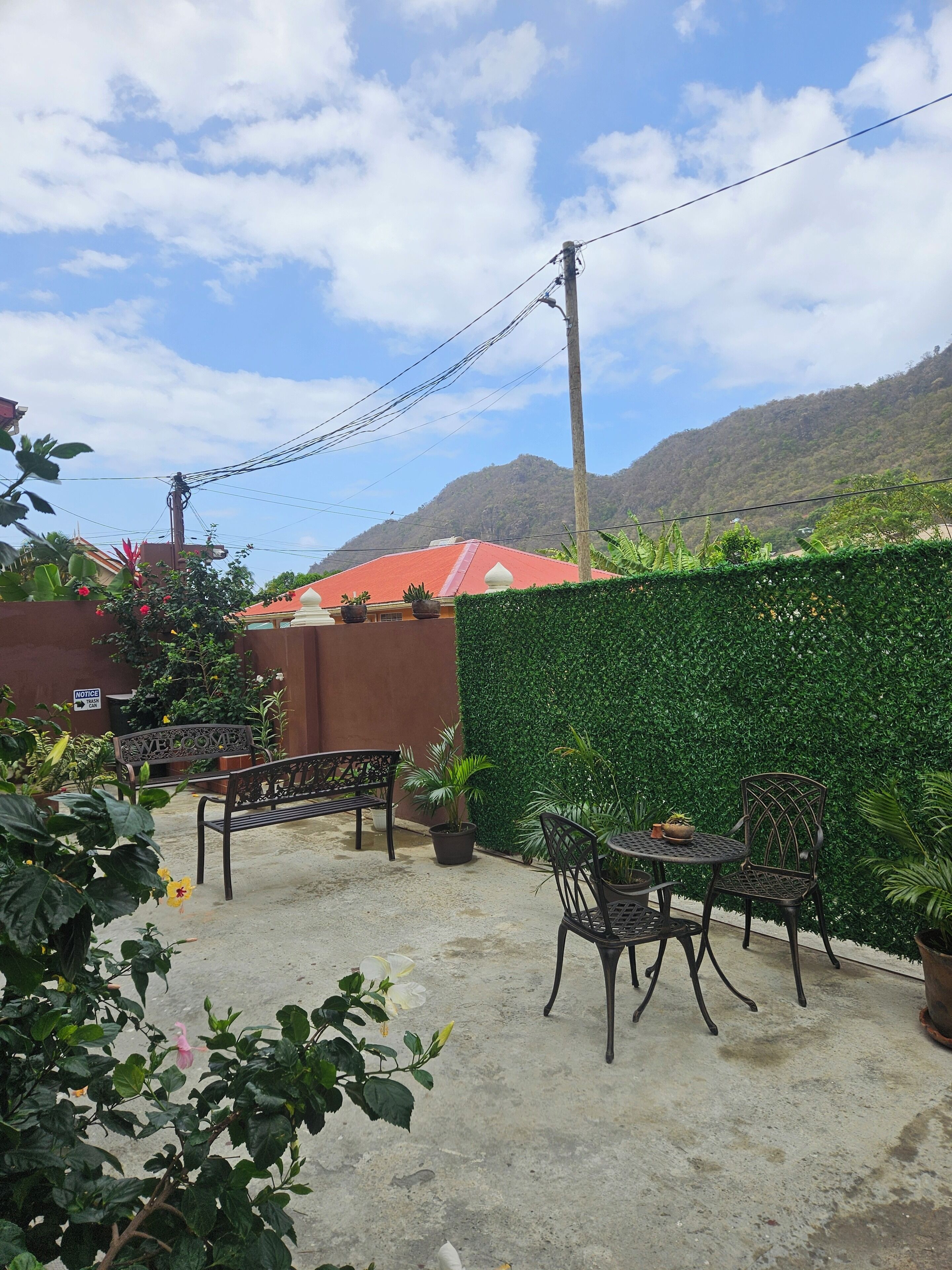 An outdoor patio with a bench and table, surrounded by potted plants and a green hedge.