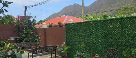 An outdoor patio with a bench and table, surrounded by potted plants and a green hedge.