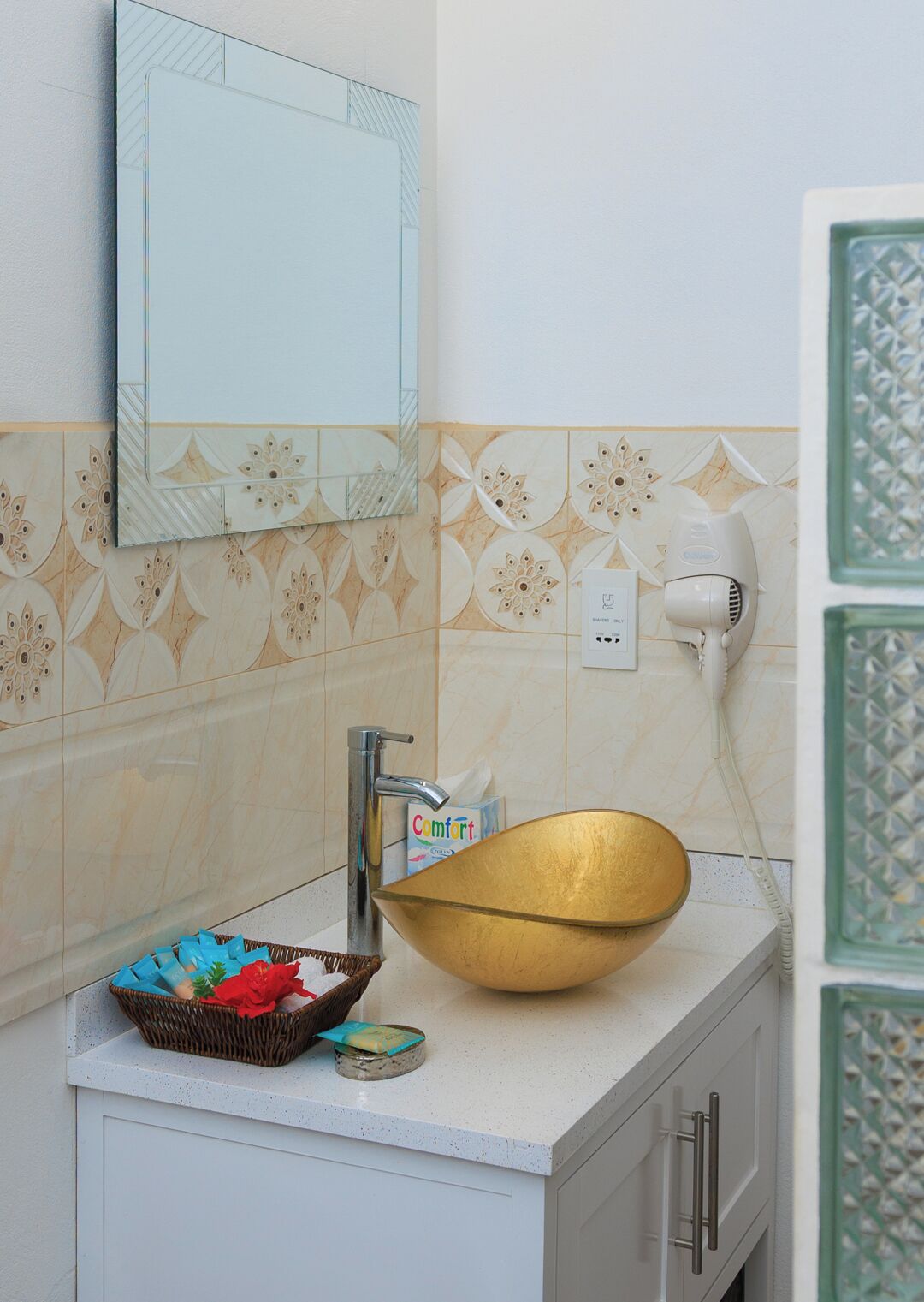 A bathroom with a large gold bowl sink, a mirror, a hairdryer, and a basket with toiletries.