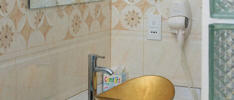 A bathroom with a large gold bowl sink, a mirror, a hairdryer, and a basket with toiletries.