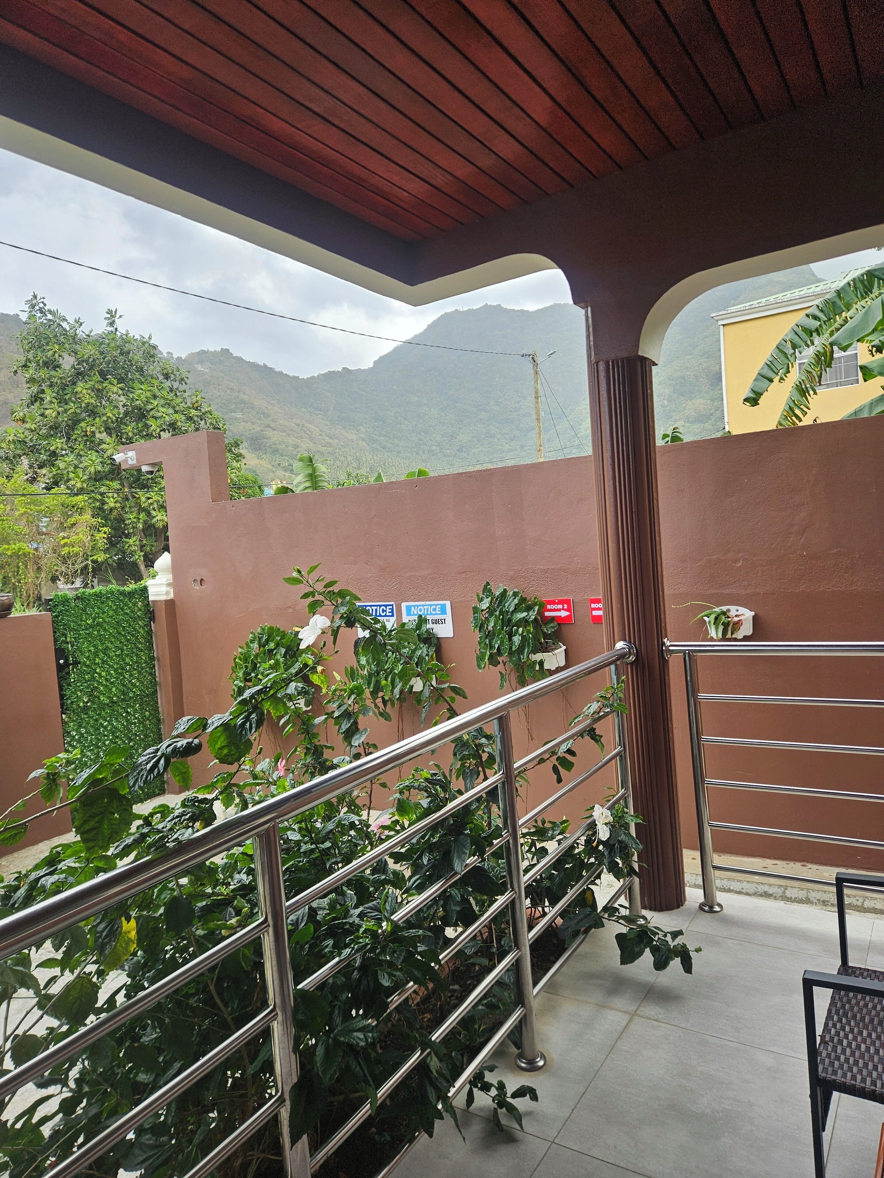 Un balcone riparato con ringhiere in metallo, vista sulle montagne e un muro marrone con cartelli che riportano 'NOTICE' e 'ROOM 2'.