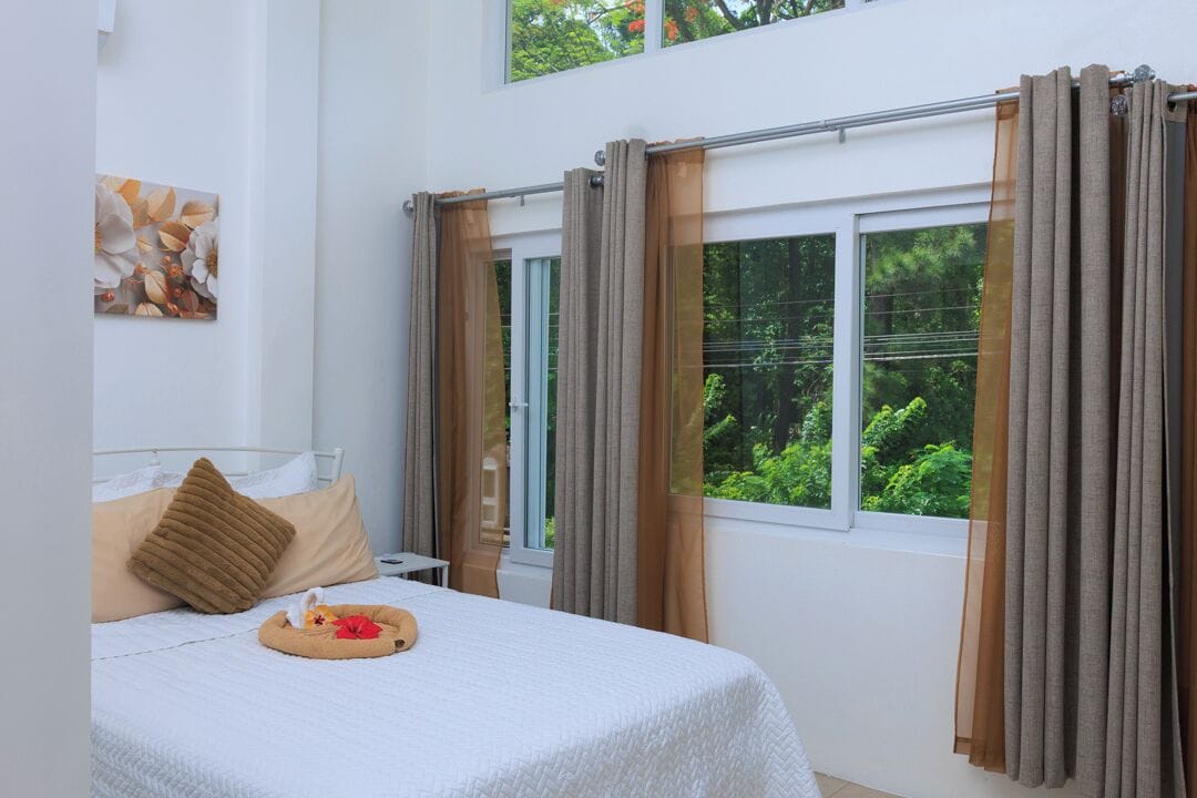 Une chambre à coucher avec un lit, une fenêtre avec des rideaux, un tableau au mur et une vue sur la verdure à l’extérieur.