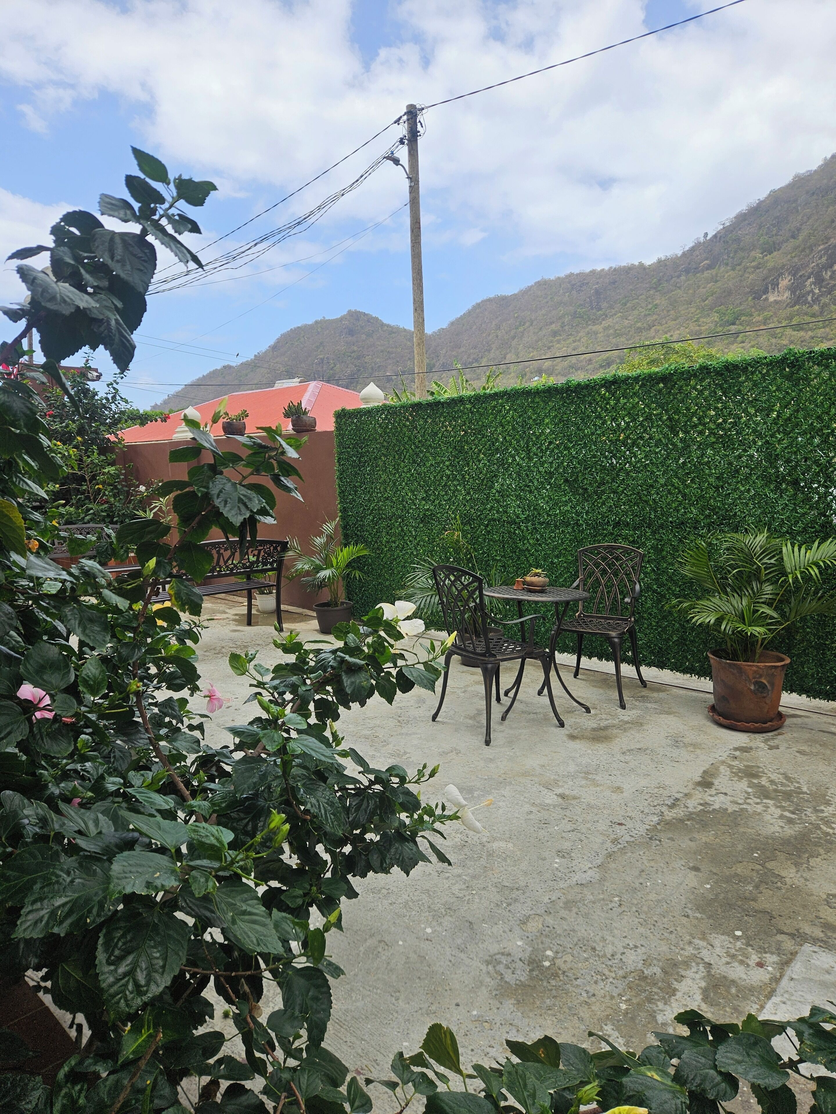 Un patio al aire libre con una mesa y sillas, rodeado de abundante vegetación y un edificio rojo al fondo.