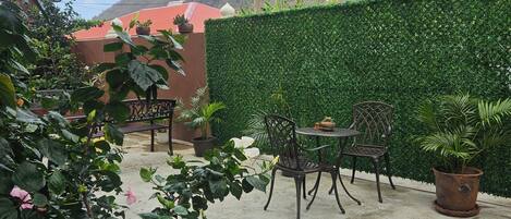 Un patio al aire libre con una mesa y sillas, rodeado de abundante vegetación y un edificio rojo al fondo.