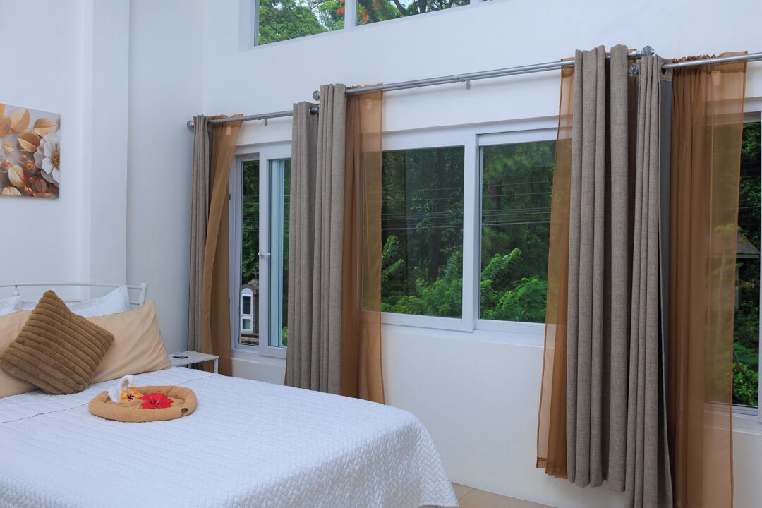 A bedroom with a bed, a bedside table, a window with curtains, and a view of greenery outside.