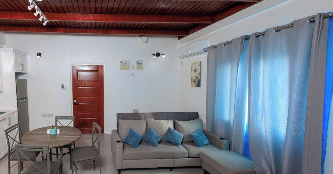 A living room with a sofa, a dining table with chairs, a wooden ceiling, and a door.