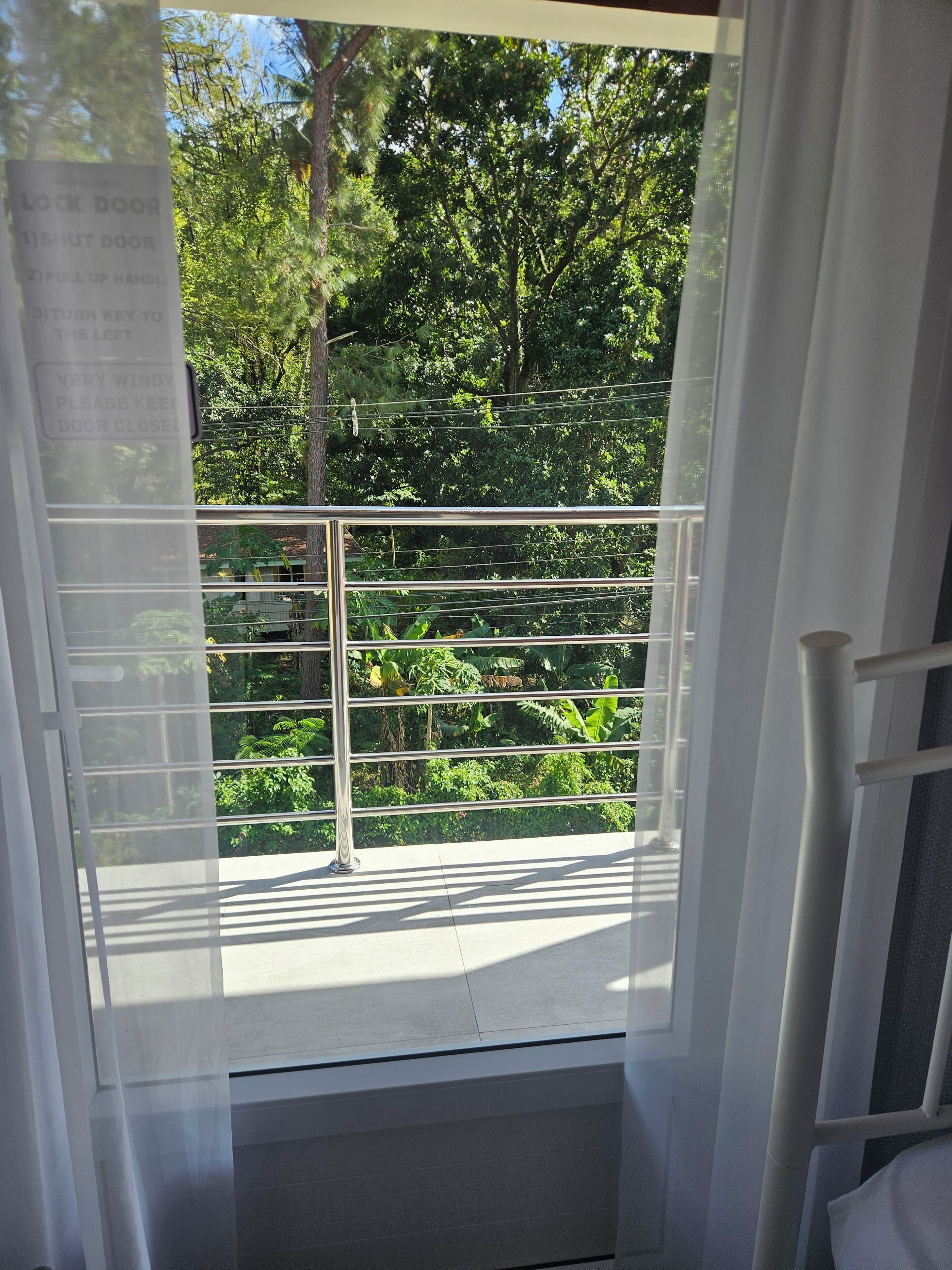 A window with horizontal blinds, a metal railing, and a view of greenery outside.