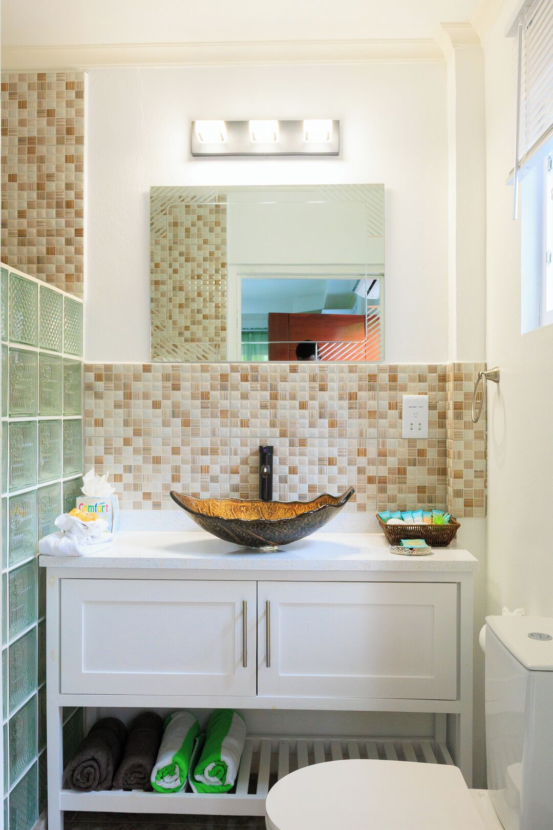 A bathroom with a mosaic tile backsplash, a large mirror, a vessel sink, a toilet, and a cabinet with towels.