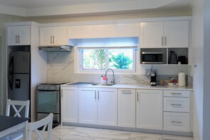 A kitchen with white cabinets, a stove, a sink, a window with a view of greenery, and a microwave.