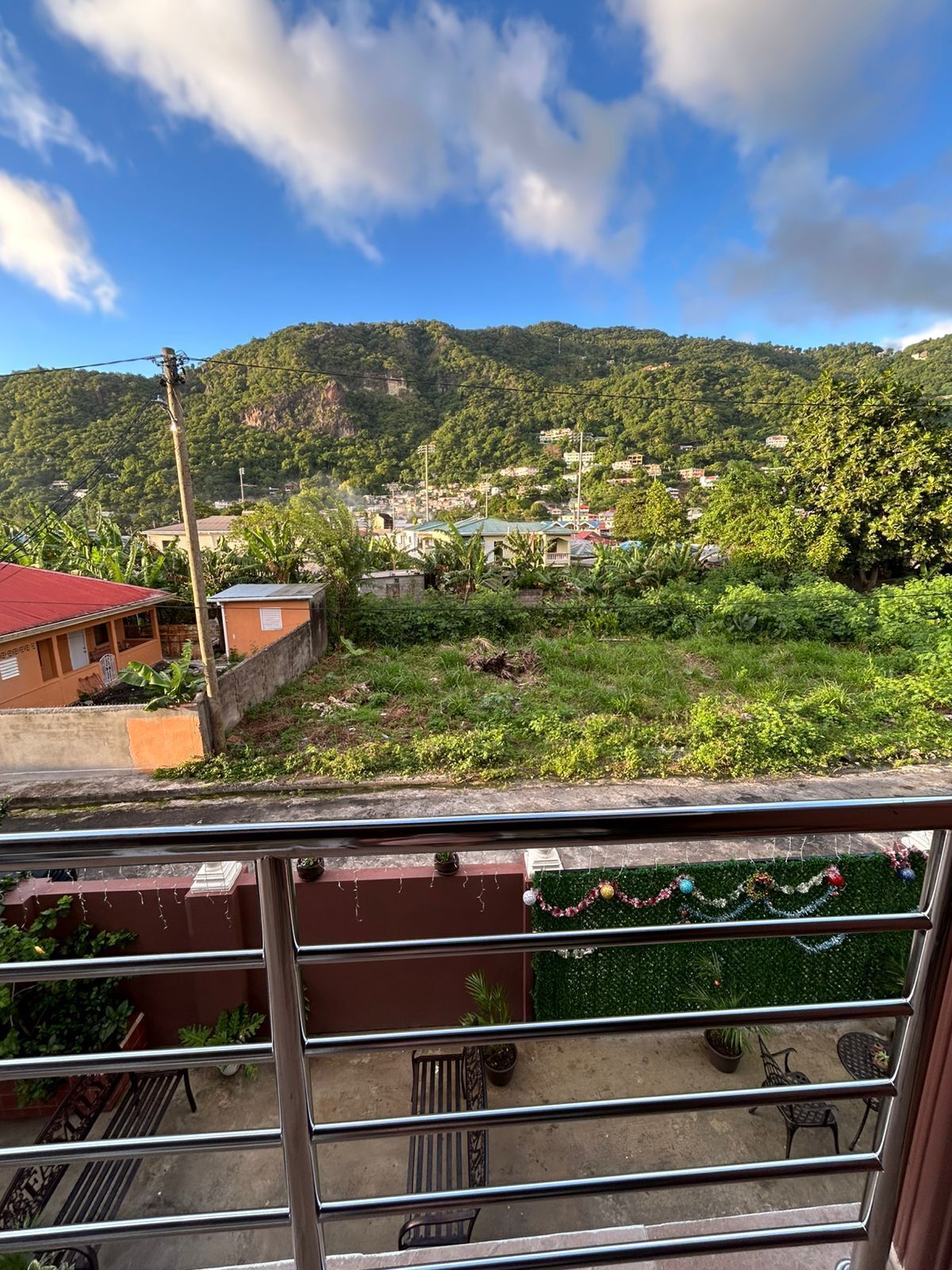 Un balcón con vistas a un pueblo en la ladera, un edificio de techo rojo y un césped verde con una guirnalda decorativa.
