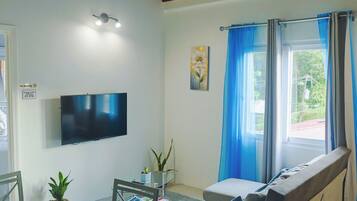 Una sala de estar con una televisión de pantalla plana, un sofá, una mesa de comedor con sillas y una ventana con cortinas azules.