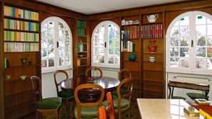Een bibliotheek met houten boekenkasten, een ronde tafel met stoelen en rondbogenramen.