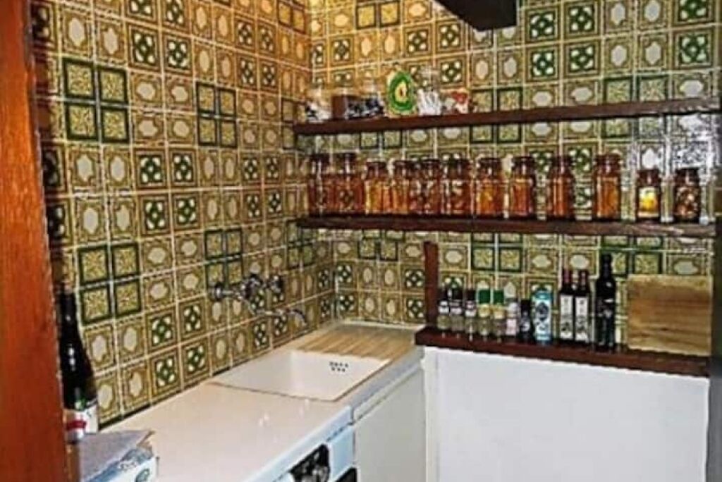 Una cocina con salpicadero de azulejos estampados, un lavabo y estantes con frascos y botellas.