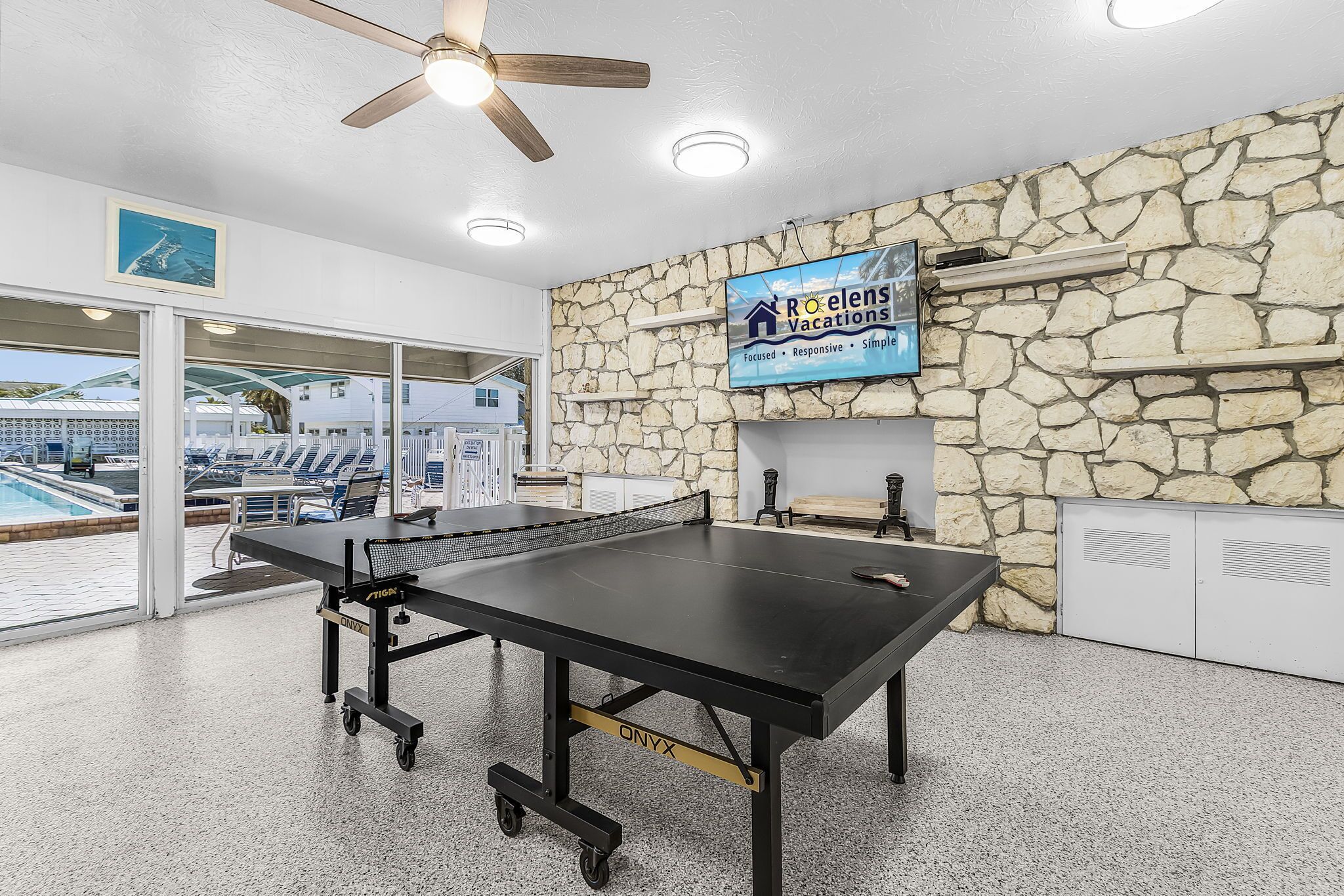 A ping pong table in a room with a stone wall, a ceiling fan, and a sign for Roelens Vacations.