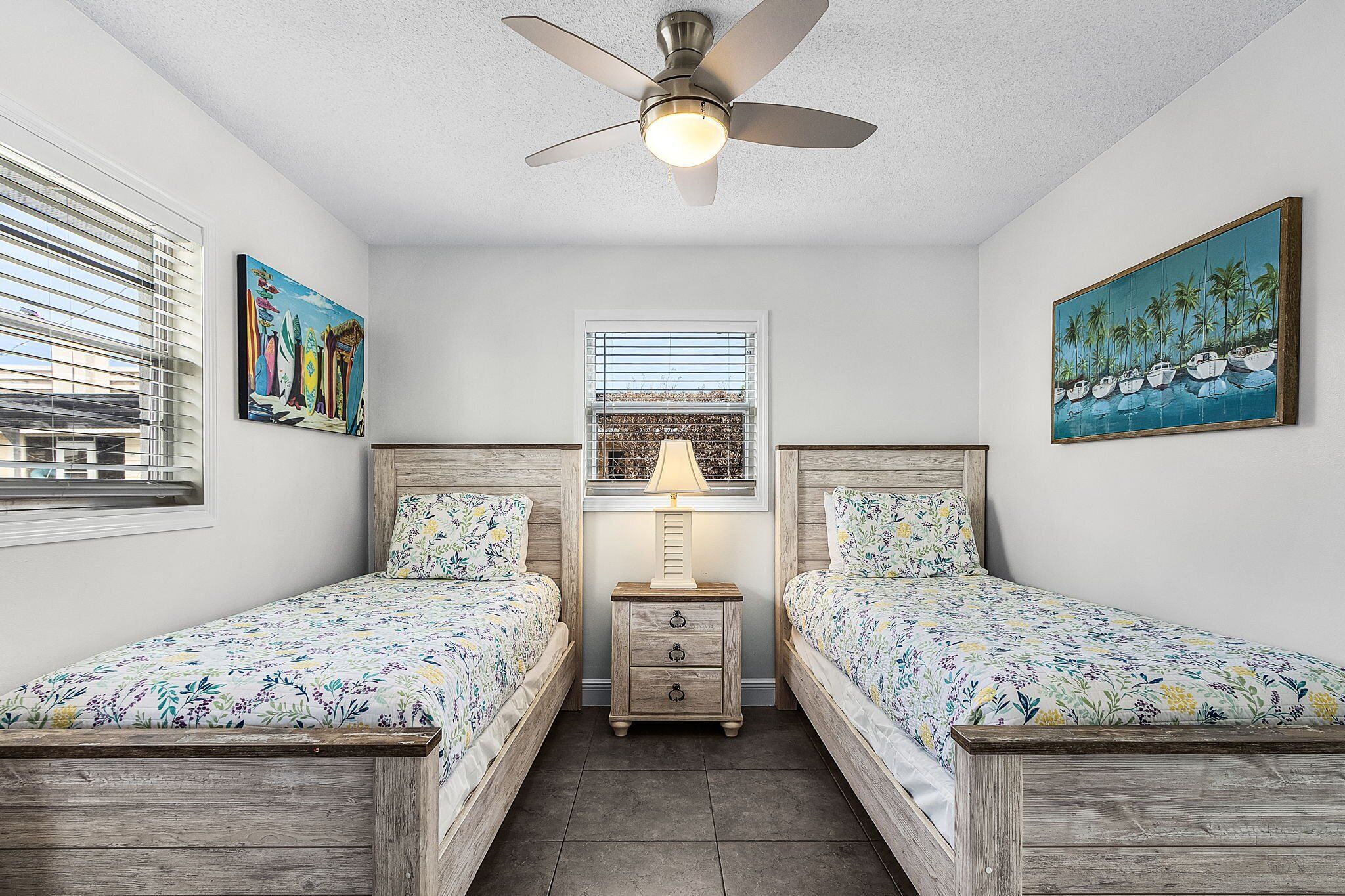 A bedroom with two wooden beds, a ceiling fan, a nightstand with a lamp, and two paintings on the walls.