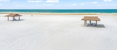A beach with two wooden gazebos, one with a person sitting and another with a person standing.