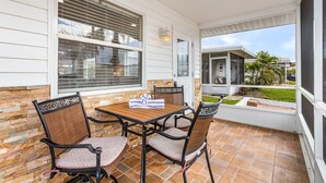 A covered patio with a table and chairs, large windows, and a view of a house and greenery. - A Great Day At The Beach! - Aloha Kai #6 - Roelens (Sarasota)