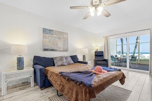 A bedroom with a bed, two bedside tables, a ceiling fan, a large window with a view of the beach, and a painting on the wall. - Beachfront - Family-Friendly Beach Escape with Pool, Porch & Sun-Filled Fun – Aloha Kai #24 (Siesta Key)