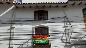 A building with a white facade, a balcony with a flag, and a wooden door. - Krass casa de huespedes (Sucre)