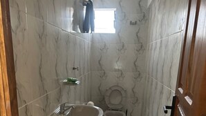 A small bathroom with white marble walls and a wooden door. It includes a sink, a toilet, and a window. - Krass casa de huespedes (Sucre)
