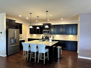 A modern kitchen with dark cabinetry, stainless steel appliances, a central island with bar stools, and pendant lighting.