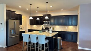 A modern kitchen with dark cabinetry, stainless steel appliances, a granite countertop, and a central island with bar stools. - Spacious LUXE Home in RTP / near Airport (Durham)
