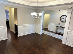 A spacious room with hardwood flooring, white wainscoting, a chandelier, and a clock on the wall.