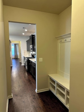 A hallway with a dark wooden floor, a white bench, and a kitchen area with dark cabinets and a white countertop. - Spacious LUXE Home | RTP/Airport (Durham)