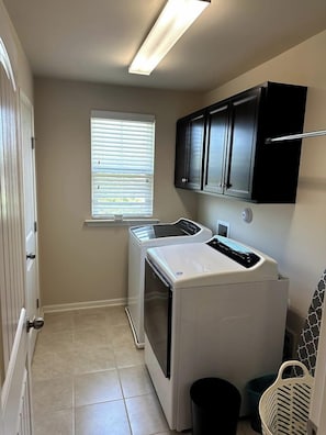 A laundry room with a washing machine, a dryer, a window with blinds, and a trash can. - Spacious LUXE Home | RTP/Airport (Durham)