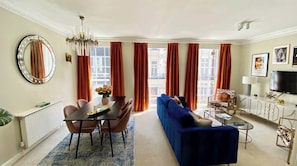 A modern living room with a blue sofa, a dining table with chairs, a chandelier, and large windows with orange curtains.