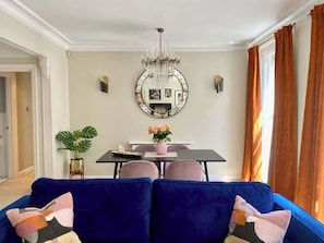 A living room with a blue sofa, a dining table with chairs, a chandelier, and a large mirror on the wall.