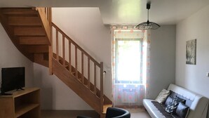 A living room with a wooden staircase, a white sofa, a black armchair, and a TV on a wooden stand. - GITE BROCELIANDE - CARENTOIR (CARENTOIR)