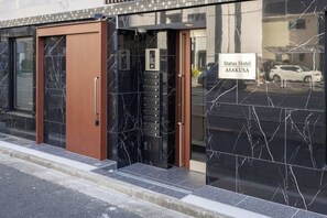 L’entrée d’un hôtel, avec une enseigne indiquant « Status Hotel ASAKUSA », une porte en bois, un ascenseur en verre et une façade en marbre noir.