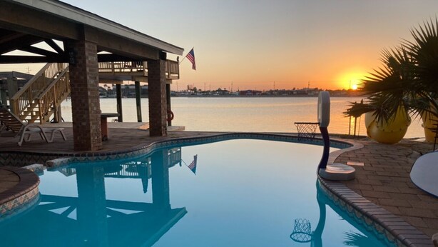 En swimmingpool med udsigt til solnedgang, en overdækket terrasse og et flag.