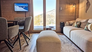 A cozy living room with a sofa, ottoman, and chairs, a television mounted on the wall, and a large window offering a scenic view. - Beauregard 1 by Interhome (Nendaz)