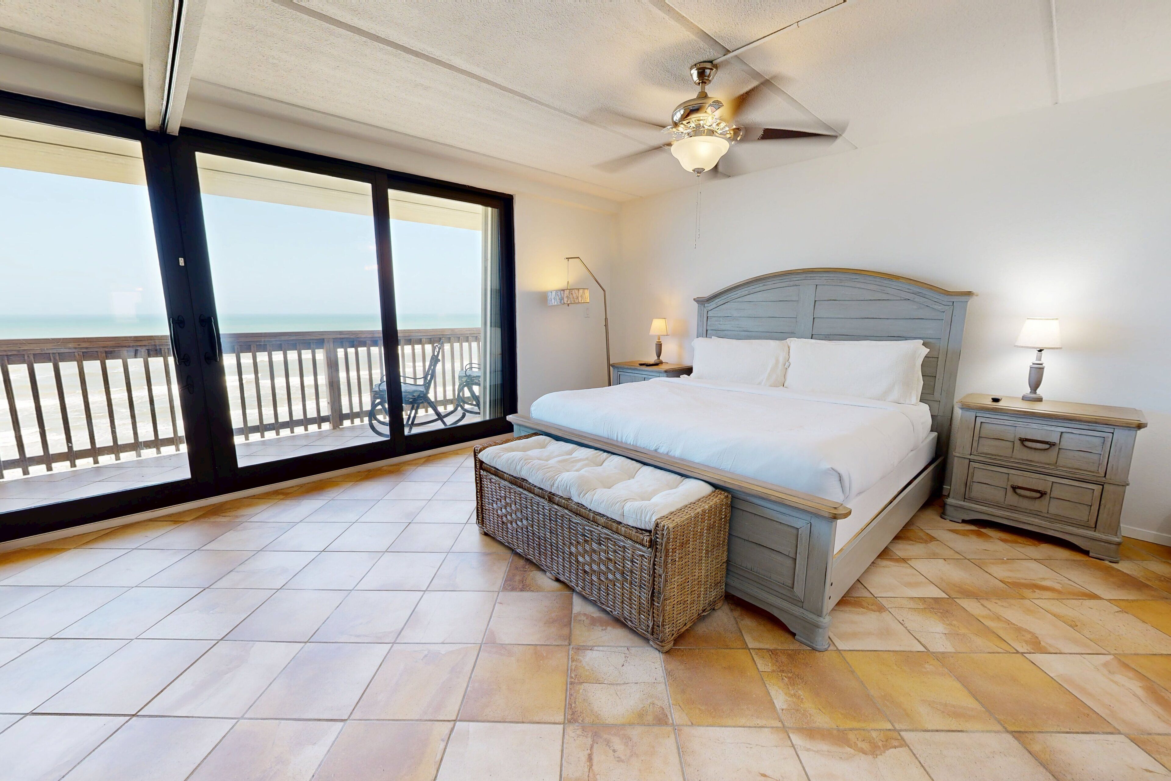 A bedroom with a large bed, a ceiling fan, a nightstand, a wicker bench, and a view of the beach through a sliding door.