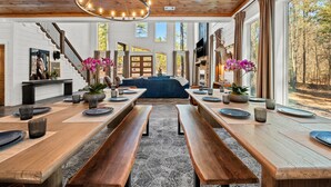 Un vaste espace dédié à la salle à manger, avec des tables et des bancs en bois, de grandes fenêtres donnant sur la forêt et un lustre suspendu au plafond.
