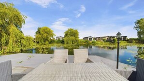 A serene outdoor area with a paved deck, wicker chairs, and a calm water body surrounded by lush greenery and modern buildings in the background. - Lakeside Lodge Retreat with Views (Fenstanton)