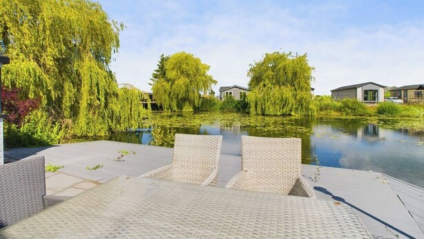 A serene outdoor area with a paved deck, wicker chairs, and a calm water body surrounded by lush greenery and modern buildings. - Lakeside Lodge Retreat with Views (Fenstanton)