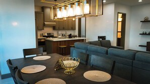 Uma cozinha moderna com uma ilha central, uma sala de jantar com mesa de madeira escura e jogos americanos trançados, e um sofá cinza na sala de estar.