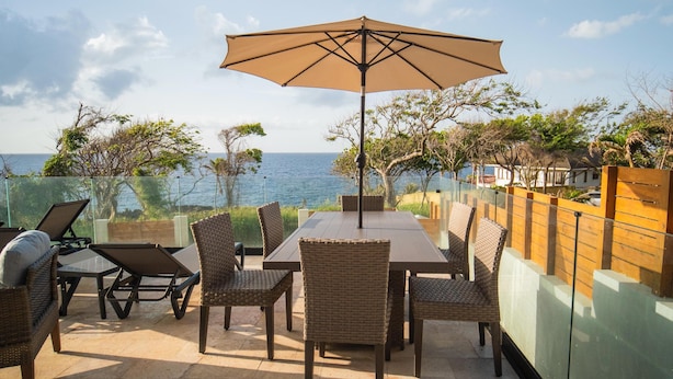 Une terrasse avec une table et des chaises, un grand parasol et une vue sur l’océan.