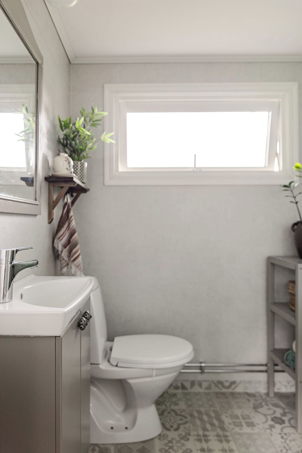 Une salle de bain avec un lavabo blanc, des toilettes, une fenêtre et une étagère avec des plantes.