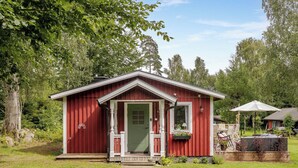 A small red wooden cabin with a green door, a porch, and a small garden area with plants and a bench.
