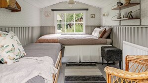 A cozy bedroom with a sloped ceiling, a large window, a bed with a patterned bedspread, a small bench, and a wicker chair.