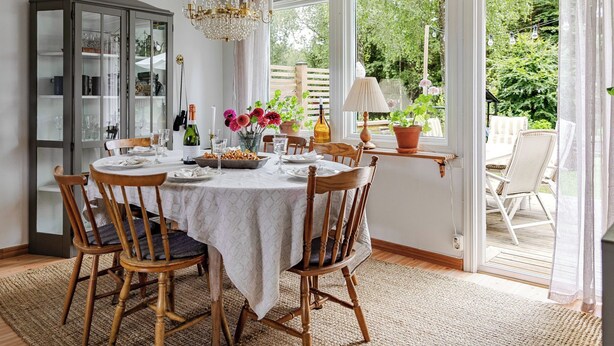 A dining area with a table set for four, surrounded by wooden chairs. A chandelier hangs above, and a window offers a view of greenery outside.