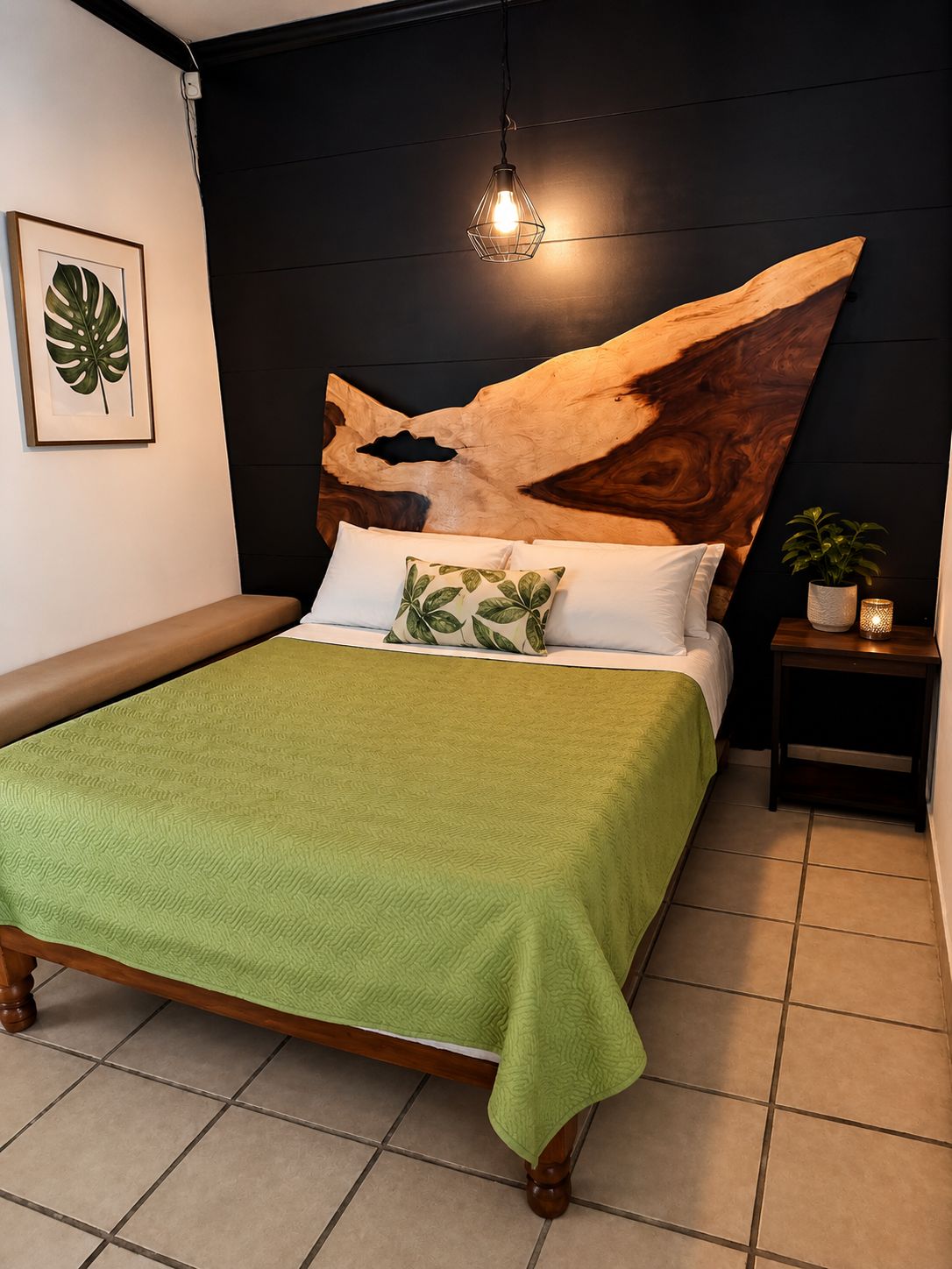 A bed with a green blanket, a wooden headboard with a leaf design, a bedside table with a plant, and a wall-mounted light fixture.