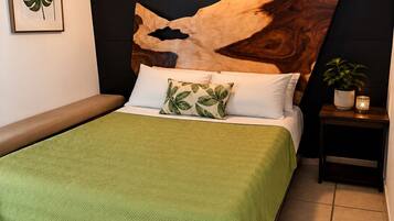 A bed with a green blanket, a wooden headboard with a leaf design, a bedside table with a plant, and a wall-mounted light fixture.