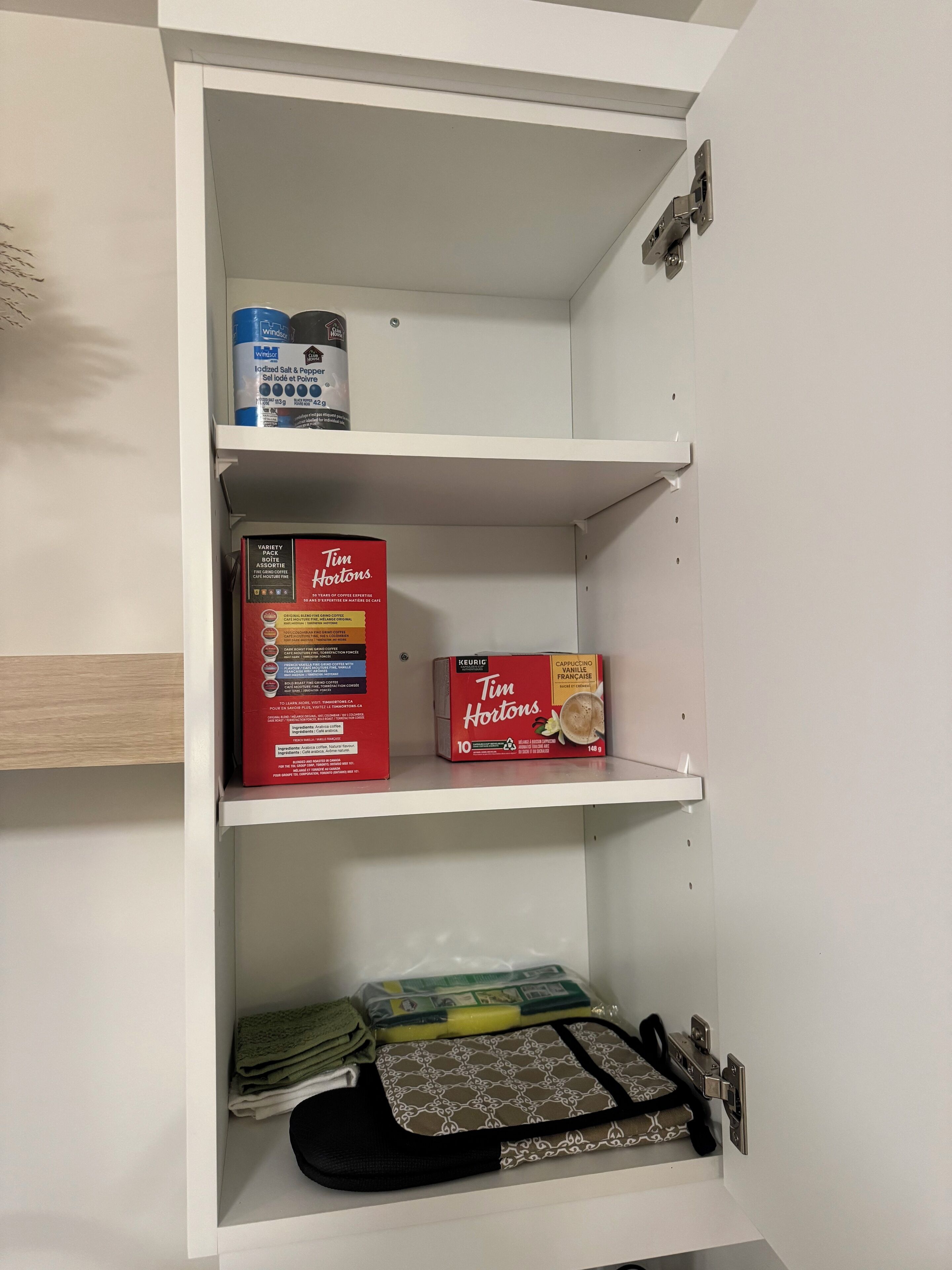 Un meuble blanc avec des étagères contenant une boîte de café Tim Hortons, une boîte de dosettes de café Keurig et un article plié muni d’une sangle noire.